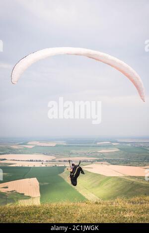 Ein Mann Gleitschirm, der vom Rand des Berges mit Feldern im Hintergrund abheben. Gleitschirmfliegen Stockfoto