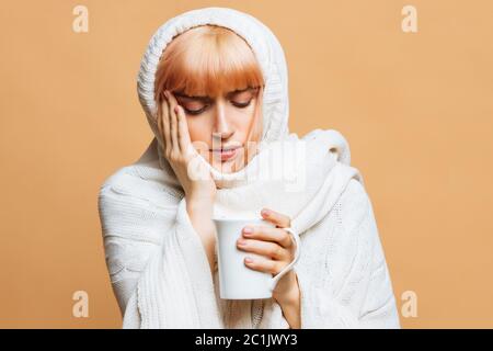 Sickness nette junge Frau mit Erdbeer blonden Haaren, warm karierten mit Kopfschmerzen, hält Tasse heißen Tee, berühren ihren Tempel, Gefühl der ersten Symptome Stockfoto