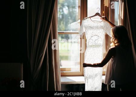Blonde Frau mit lockigen Haaren halten und Blick auf schöne weiße Hochzeitskleid auf einem Kleiderbügel, in der Nähe des Fensters in ihrem Haus, Rückansicht, Taglig Stockfoto