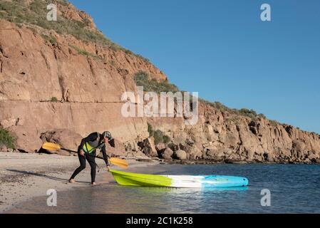 Eine Frau, die einen Neoprenanzug trägt und ein Paddel hält, macht sich bereit, auf einem Platz auf dem Kajak auf Carmen Island, Loreto, Baja California zu springen. Stockfoto