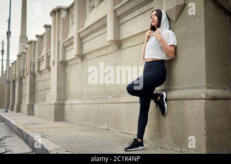 Die ganze Jogginglänge blickt weg, während sie auf dem Bürgersteig gegen die Wand in der Stadt steht Stockfoto