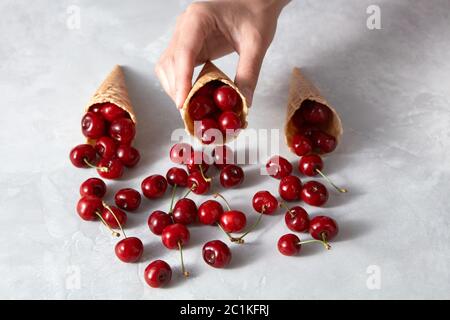 Die Hand der Frau nimmt knusprige Waffelbecher mit reife rote Kirschen auf einem grauen Stein. Stockfoto