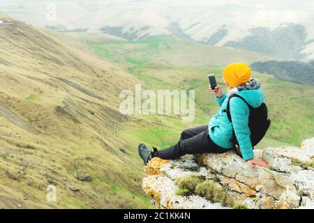 Rückansicht einer Frau, die in Winterkleidung auf einem Felsen sitzt und einen Hut auf einem Smartphone trägt. Kaukasus Stockfoto