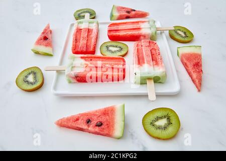 Scheiben Wassermelone und Kiwi in Form eines Kreises und eines Dreiecks um einen Teller mit einem gesunden Eis Lolly in der Form Stockfoto