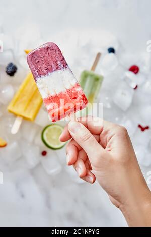 Hausgemachte gefrorene Smoothies auf einem Stock, wobei eine weibliche Hand vor einem Hintergrund von Eiswürfeln mit verschiedenen Beeren und Eis gehalten wird Stockfoto