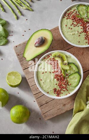 Draufsicht auf einen grünen Smoothie aus Avacado, Gurke, Kiwi, Spargel mit Leinsamen und Limettenscheiben in einer weißen Schüssel auf einem Holzkochfeld Stockfoto