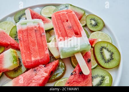 Sommerdessert aus gefrorenem Fruchtsaft Lolly mit Eis und Stücken von Wassermelone, Kiwi und Limette in einem Teller auf einem grauen Marmor-Backgr Stockfoto