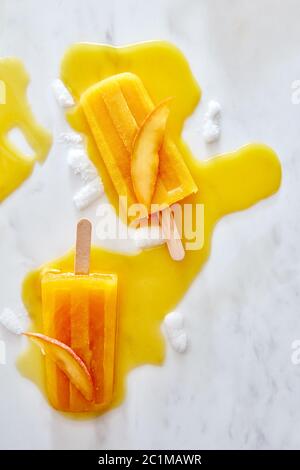 Erfrischendes Mango-Eis mit Pfirsich-Scheibe auf Holzstab auf Marmorboden, flach gelegt Stockfoto