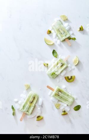Grünes Eis auf einem Stock mit einem Stück frischer Kiwi, Minzblätter und Eis, präsentiert in Form eines Eckrahmens auf einem grauen ma Stockfoto