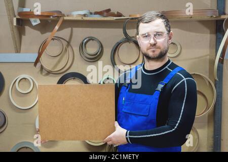 Der Hersteller von Möbeln in Spezialkleidung und Brille hält am Arbeitsplatz eine leere Tafel für den Text. Handwerk p Stockfoto