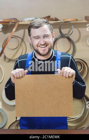 Der Hersteller von Möbeln in Spezialkleidung und Brille hält am Arbeitsplatz eine leere Tafel für den Text. Handwerk p Stockfoto