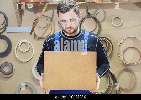 Der Hersteller von Möbeln in Spezialkleidung und Brille hält am Arbeitsplatz eine leere Tafel für den Text. Handwerk p Stockfoto