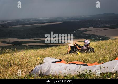 Ein professioneller Gleitschirm in voller Ausrüstung und ein Helm liegt und ruht auf dem Gras hoch in den Bergen und schaut auf die Wolken Stockfoto