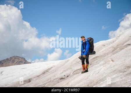 Ein Bergsteiger mit Rucksack geht im Rollstuhl, steht auf einem staubigen Gletscher mit Sneakers in den Händen zwischen Rissen in der m Stockfoto