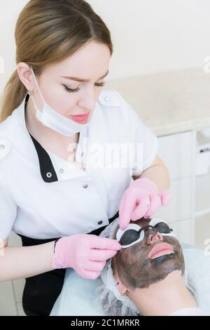 Der Kosmetologe in rosa Handschuhen mit Pinsel trägt eine Carbon-Maske zum Peeling auf das Gesicht eines jungen Mädchens in einem Kosmetologie-Roo Stockfoto