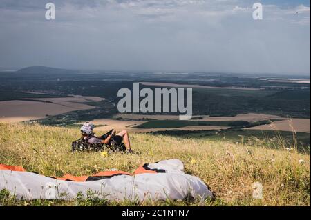 Ein professioneller Gleitschirm in voller Ausrüstung und ein Helm liegt und ruht auf dem Gras hoch in den Bergen und schaut auf die Wolken Stockfoto