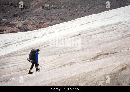 Ein Bergsteiger mit Rucksack geht in Steigeisen entlang eines staubigen Gletschers mit Gehwegen in den Händen zwischen Rissen in der Stockfoto