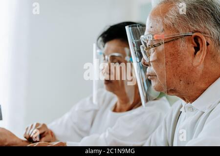 Seitenansicht eines älteren asiatischen Mannes und einer älteren Frau in Gesichtsschilden, die während einer Coronavirus-Pandemie im hellen Raum ruhen Stockfoto