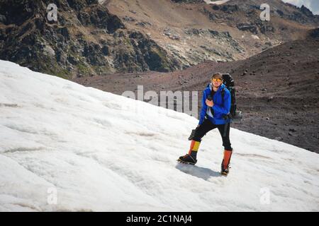 Ein Bergsteiger mit Rucksack geht im Rollstuhl, steht auf einem staubigen Gletscher mit Sneakers in den Händen zwischen Rissen in der m Stockfoto