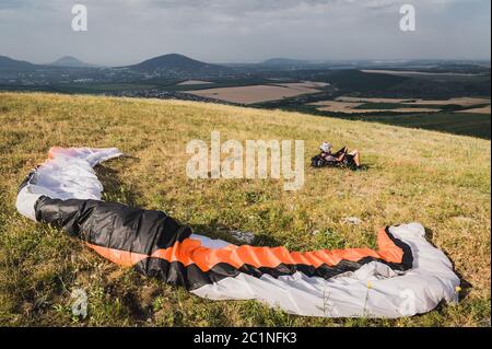 Ein professioneller Gleitschirm in voller Ausrüstung und ein Helm liegt und ruht auf dem Gras hoch in den Bergen und schaut auf die Wolken Stockfoto