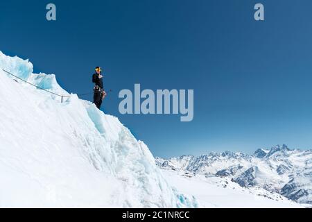 Der Bergsteiger steht am Rand des Gletschers mit einer Schneeschaufel in den Händen und zeigt Shaks Geste gegen den blauen Himmel Stockfoto