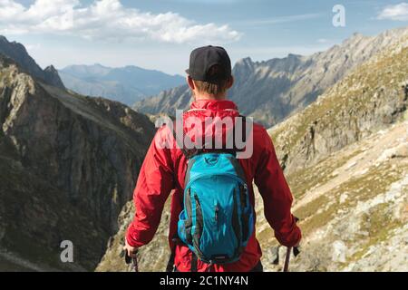 Ein bärtiger Mann in Sonnenbrille und einer Mütze mit Rucksack steht auf einem Felsen und blickt in ein felsiges Tal hoch im Berg Stockfoto