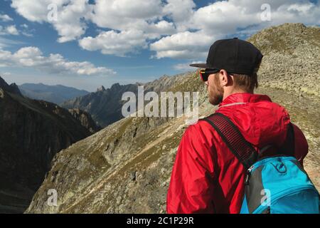 Ein bärtiger Mann in Sonnenbrille und einer Mütze mit Rucksack steht auf einem Felsen und blickt in ein felsiges Tal hoch im Berg Stockfoto