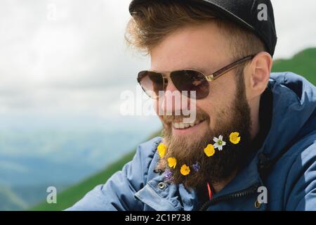 Nahaufnahme eines fröhlichen bärtigen Mannes in Sonnenbrille und einer grauen Mütze mit Wildblumen im Bart. Weiche Brutalität und gute ma Stockfoto