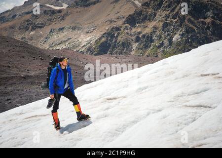 Ein Bergsteiger mit Rucksack geht im Rollstuhl, steht auf einem staubigen Gletscher mit Sneakers in den Händen zwischen Rissen in der m Stockfoto