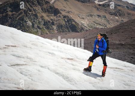 Ein Bergsteiger mit Rucksack geht im Rollstuhl, steht auf einem staubigen Gletscher mit Sneakers in den Händen zwischen Rissen in der m Stockfoto