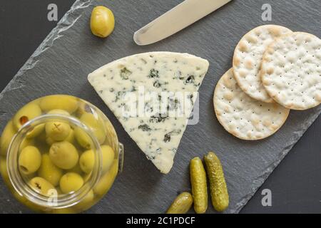 Blauer Käsekeil mit Crackern und Glas grüner Oliven auf schwarzem Schiefer Käseplatte isoliert Stockfoto