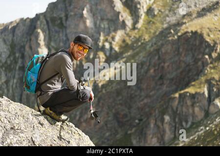 Ein bärtiger Hipster-Tourist in Sonnenbrille mit Rucksack sitzt am Rande einer Klippe hoch in den Bergen nahe dem Berg Stockfoto