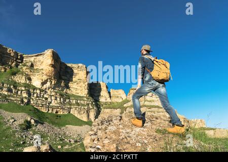 Stilvoller bärtiger männlicher Reisender in Sonnenbrille und einer Mütze mit Rucksack in einem Denim-Anzug und gelben Schuhen steht mit dem Rücken auf einem Stockfoto