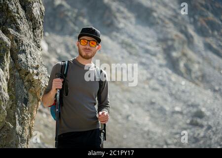 Portier eines stylischen Hipster-Reisenden mit Bart und Rucksack in Sonnenbrille und Mütze mit Trekkingstöcken steht auf einer Rock ag Stockfoto