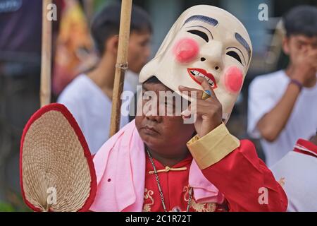 Eine Prozession während des vegetarischen Festivals in Phuket, Thailand, mit einem Mann, der seine lächelnde chinesische Maske hebt, um sein düsteres Gesicht darunter zu enthüllen Stockfoto