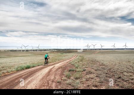 Mann auf dem Mountainbike fährt auf der Straße zum Windturbinenpark nahe dem See gegen den blau bewölkten Himmel. Ökologische und Zero Waste Lifestyle Konzepte. Stockfoto