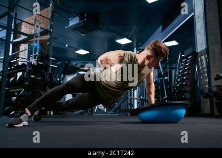 Muskulöser kaukasischer Mann, der am Boden mit Balance Halbball im Sportverein trainiert. Nahaufnahme des jungen Sportlers Muskeln bauen, tun Liegestützen auf einer Hand in leeren Fitnessraum. Konzept des Bodybuilding. Stockfoto