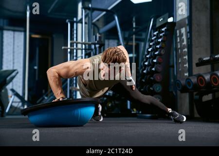 Vorderansicht des muskulösen kaukasischen Mannes Training auf dem Boden mit Balance Halbball. Nahaufnahme des jungen Sportlers Muskeln bauen, tun Liegestützen auf einer Hand in leeren Fitnessraum. Konzept des Bodybuilding. Stockfoto