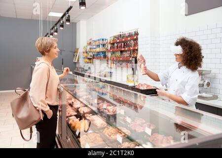 Seitenansicht der jungen lächelnden Frau mit lockigen Haaren demonstriert positive Dame rohes Schweinefleisch. Fröhliches Mädchen hinter Ladentisch mit großen Sortiment zeigt Fleisch in Schüssel aus Kühlschrank, Supermarkt. Stockfoto