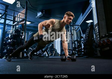 Muskulöser kaukasischer Mann, der am Boden mit Hanteln im Sportverein trainiert. Nahaufnahme des jungen Sportlers Muskeln bauen, tun Liegestützen auf einer Hand in leeren Fitnessraum. Konzept des Bodybuilding. Stockfoto