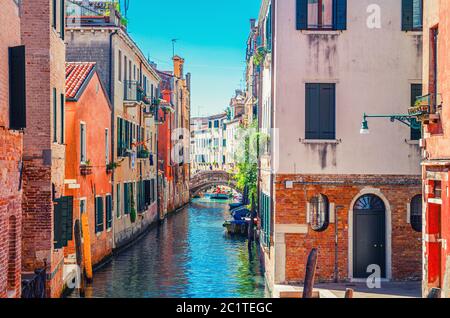 Venedig Stadtbild mit schmalem Wasserkanal mit Booten, die zwischen alten bunten Gebäuden und Steinbrücke, Venetien Region, Norditalien vertäut sind. Typisch venezianische Sicht, blauer Himmel im Hintergrund Stockfoto