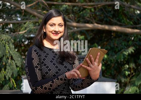 Attraktive indische Frau trägt körperbetontes Kleid steht gegen Bäume Surfen Internet auf digitalen Tablet, horizontal Medium Porträt, Kopierer Raum Stockfoto