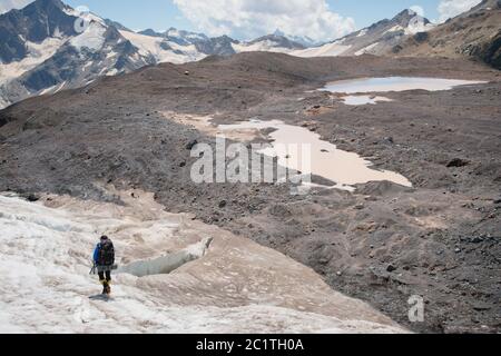 Ein Bergsteiger mit Rucksack geht in Steigeisen entlang eines staubigen Gletschers mit Gehwegen in den Händen zwischen Rissen in der Stockfoto