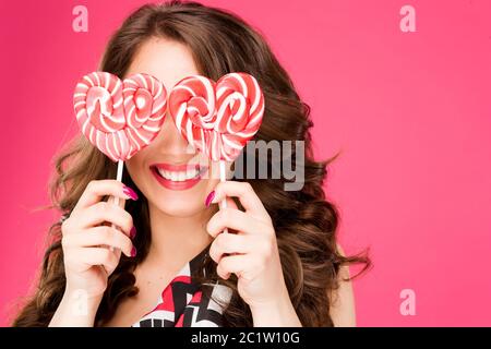 Portrait von positiven und ziemlich welligen Haaren Brünette Frau versteckt Gesicht und halten in der Hand zwei große süße gestreifte Süßigkeiten Herzen auf s Stockfoto