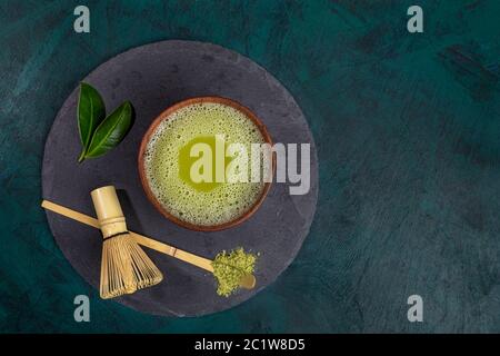 Holztasse mit grünem Matcha-Tee, Schneebesen, Löffel und Blättern auf runder Schieferplatte im smaragdgrünen Hintergrund. Matcha Tee ist Bio-asiatische gesunde Produkte Stockfoto