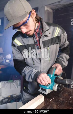 Junger Zimmermann mit Bart, der mit einem elektrischen Flugzeug mit Sägemehl-Absaugung arbeitet. Nivellierung und Schleifen von Holzstäben Stockfoto