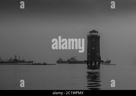 Leuchtturm und Silhouette der Fischer auf ihrem traditionellen Boot mit Industrieschiffen im Hintergrund im Sunda Kelapa Hafen, Jakarta, Indonesien Stockfoto