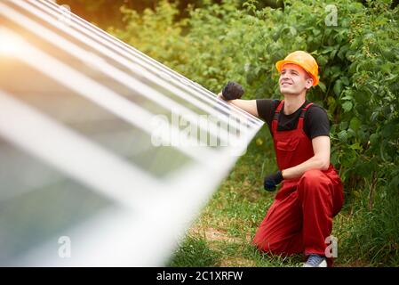 Junge Techniker in orange Uniform, Sicherheitskappe in der Nähe auf verschwommenem Hintergrund von Solarzellen und Grün. Hausbau. Alternative Energie nachhaltiges Konzept Stockfoto