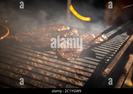 Abends ein Barbecue-Grill, auf dem leckere saftige Steaks über offenem Feuer gegrillt werden, eine Hand hält die Zange und dreht das Fleisch um. Nahaufnahme, weich Stockfoto