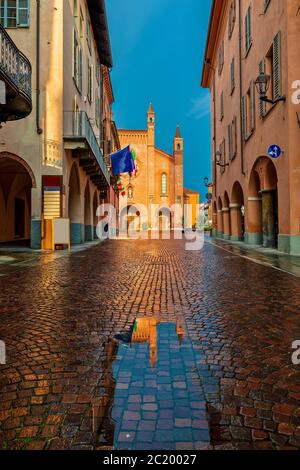 Kleine Pfütze auf schmalen Kopfsteinpflasterstraße zwischen alten Häusern und San Lorenzo Kathedrale im Hintergrund in der Stadt Alba, Piemont, Norditalien. Stockfoto